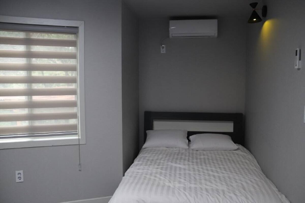 Ein Schlafzimmer mit Bett, einem Fenster mit Jalousien, einer Klimaanlage und einer Wandleuchte.