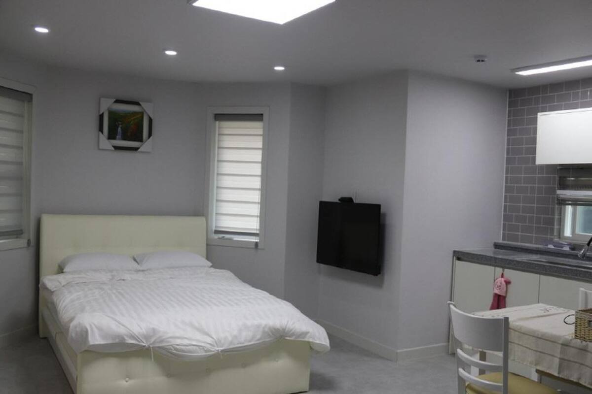 Ein Schlafzimmer mit einem Bett, einem an der Wand befestigten Fernseher, einem Fenster und einer Küchenzeile.
