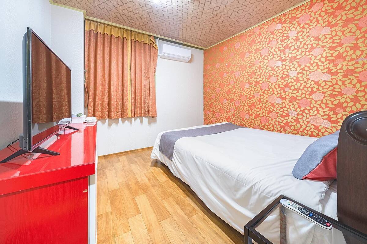 Habitación de hotel con una cama, un escritorio rojo con un televisor, una mesita con un control remoto y una pared con estampado floral.