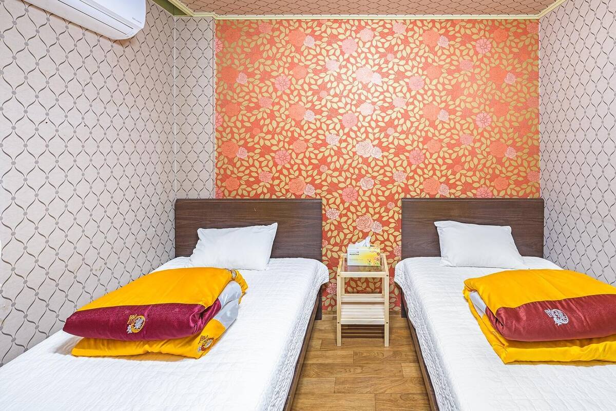 Habitación de hotel con dos camas, cada una con colchón blanco y almohada amarilla y granate. Un pequeño mesita de noche de madera con una caja de pañuelos está entre las camas. La habitación tiene papel tapiz estampado y suelo de madera.