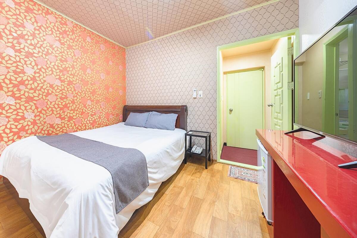 Un dormitorio con cama, una mesita de noche, un escritorio con computadora, una puerta verde y un espejo.