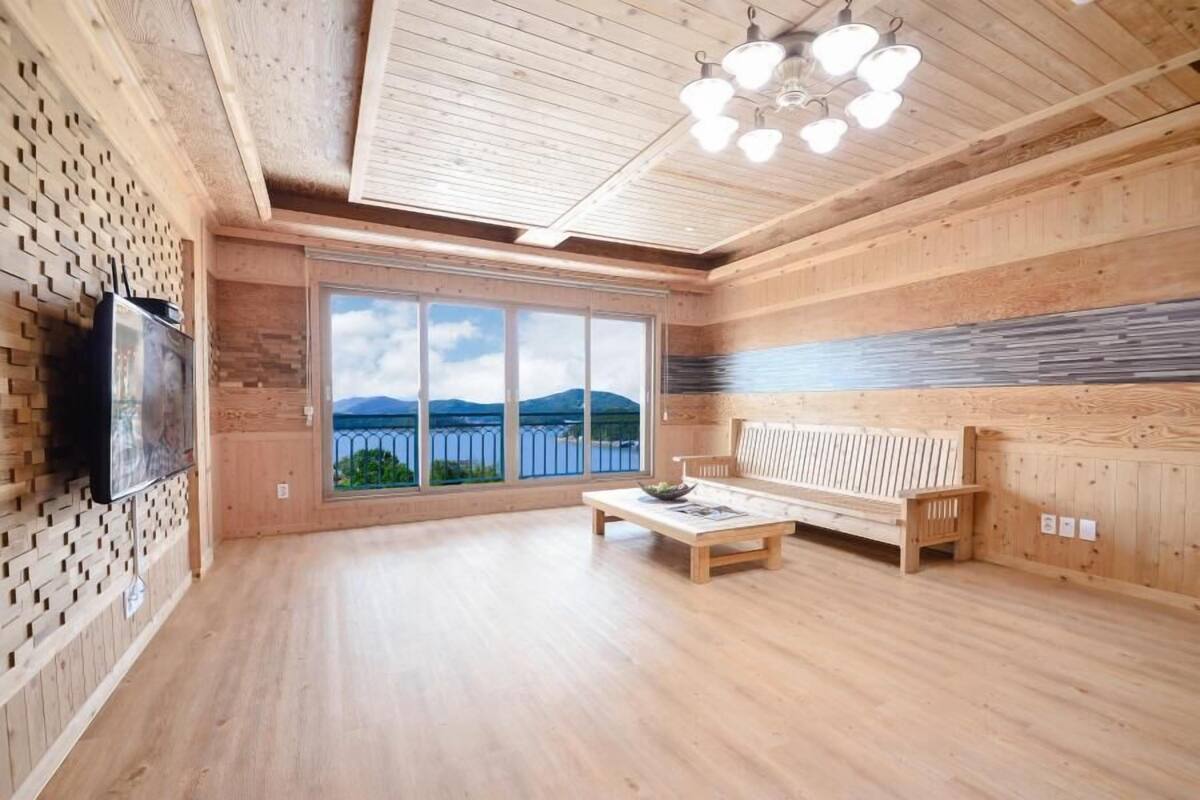Une pièce spacieuse aux murs et au sol en bois, une grande fenêtre offrant une vue sur les montagnes, un banc en bois et une table avec un bol.