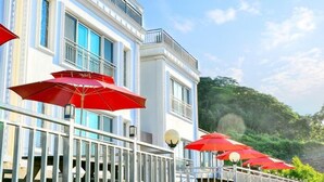 Un edificio con fachada blanca y ventanas azules, que cuenta con un balcón con sombrillas rojas y una barandilla.