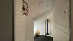 En smal hall med vit panel och svart klinkergolv. På golvet står en liten träpall och en dammsugare.