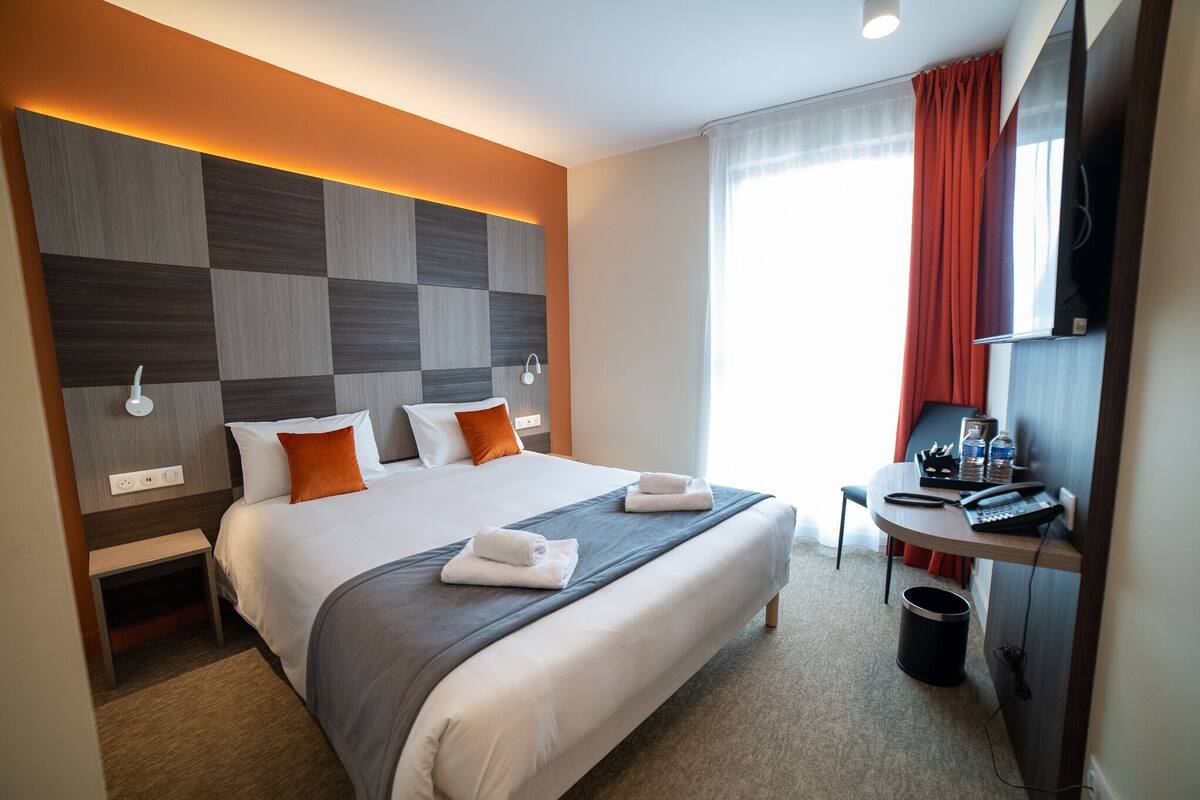 A hotel room with a bed, a desk with a computer and phone, a trash can, and a window with curtains.