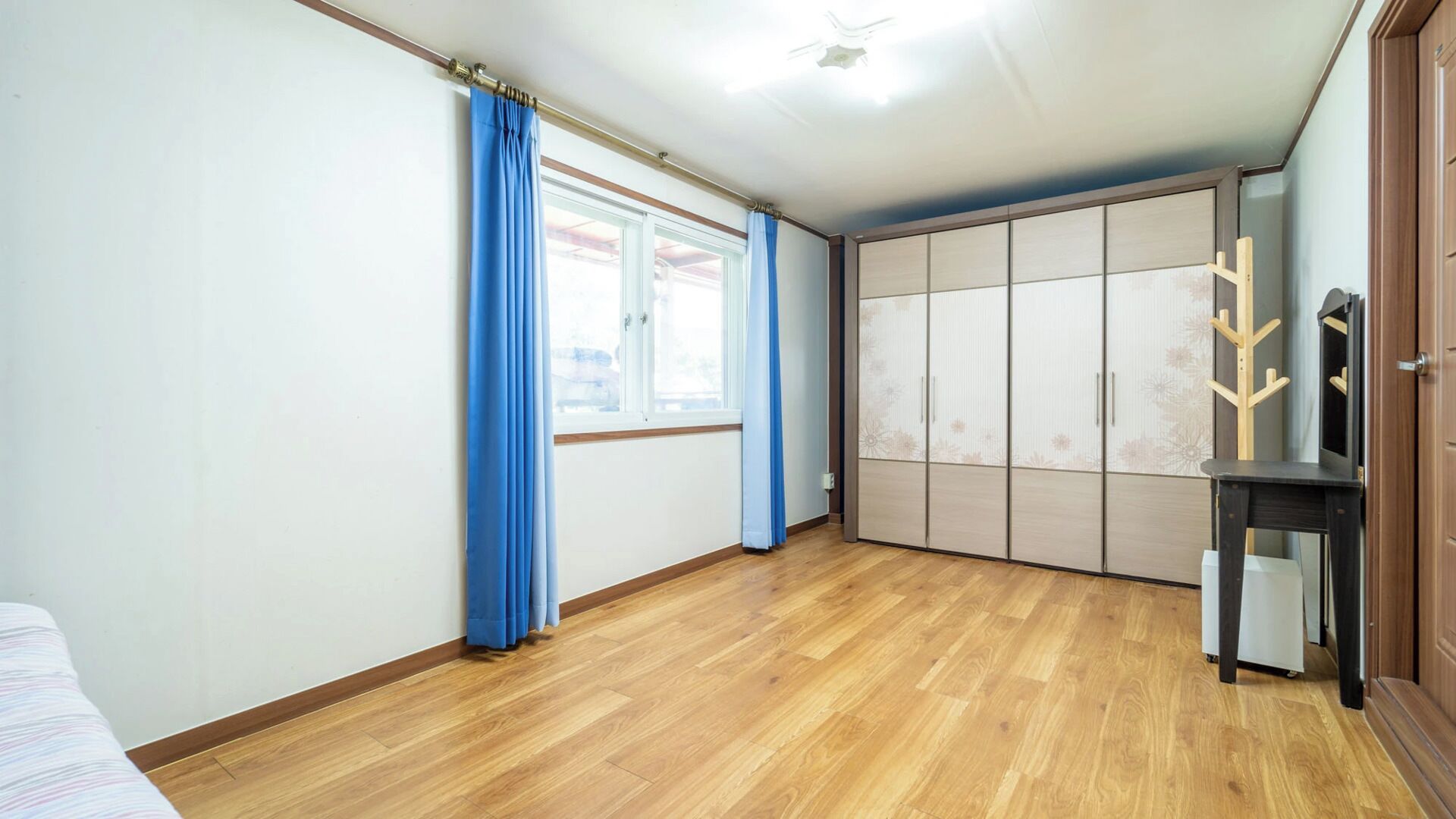 Um quarto com piso de madeira, uma janela com cortinas azuis e um guarda-roupa com várias portas.
