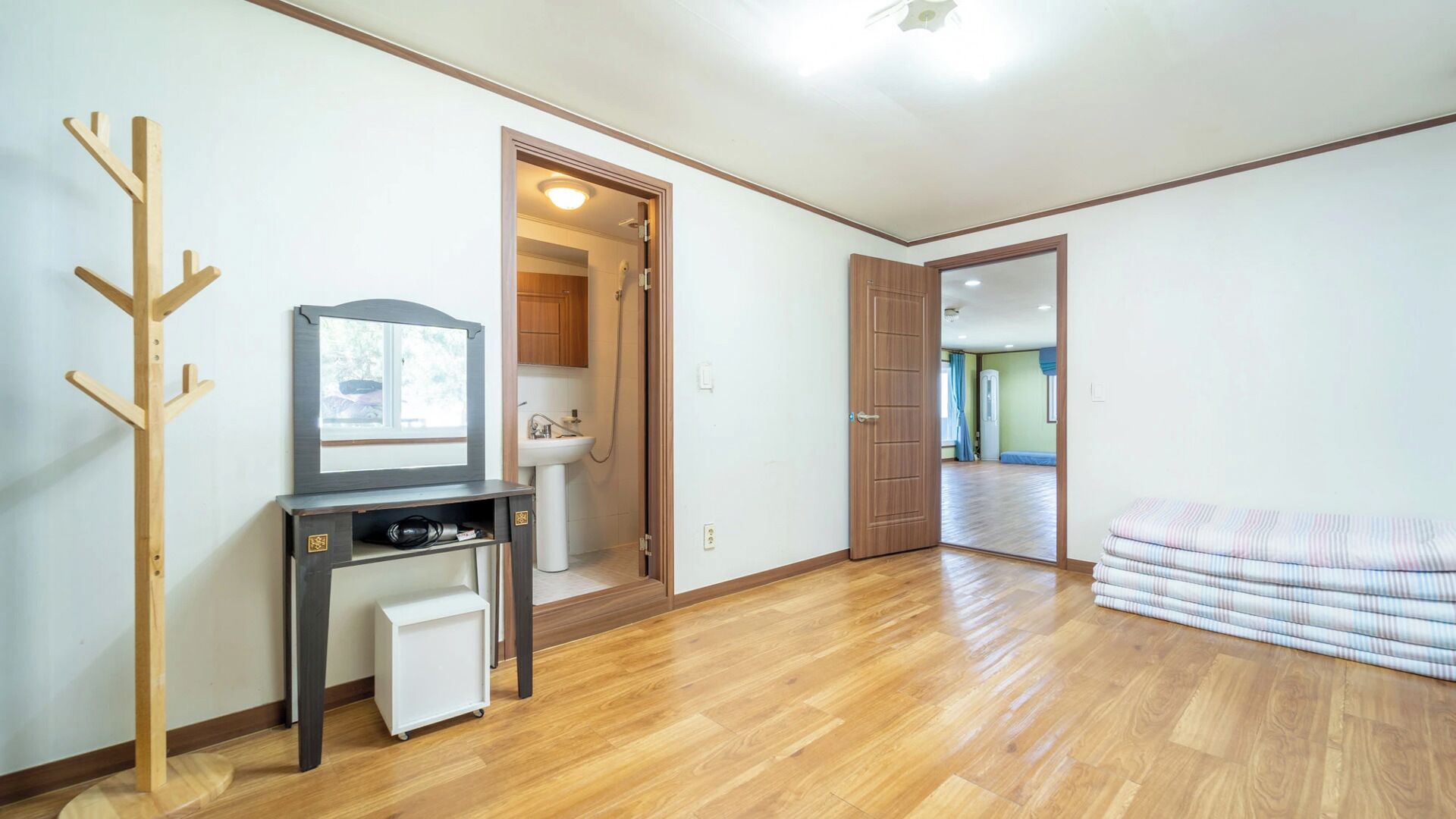 Ein Zimmer mit Holzboden, einem Kleiderständer, einem kleinen Tisch mit Spiegel, einer Toilette und einem Bett mit zusammengerollten Decken.