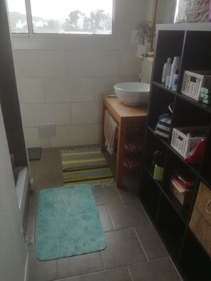 A small bathroom with a sink, a shelf with toiletries, a striped rug, and a window with a view of trees.