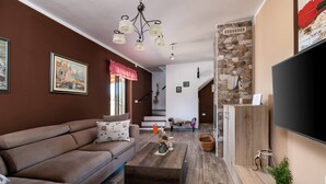 Ein Wohnzimmer mit einer Couch, einem Couchtisch und einem Flachbildfernseher. Eine Treppe führt zu einer weiteren Ebene, und eine Steinwand mit einem Bilderrahmen ist zu sehen.