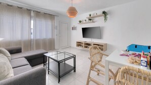 Ein modernes Wohnzimmer mit einer Couch, einem Glastisch, einem Korbstuhl, einem Fernseher und einem kleinen Tisch mit Snacks.