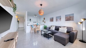 Ein modernes Wohnzimmer mit grauem Sofa, Couchtisch und TV-Möbel. Es gibt einen Essbereich mit Tisch und Stühlen, und im Hintergrund eine Küche.