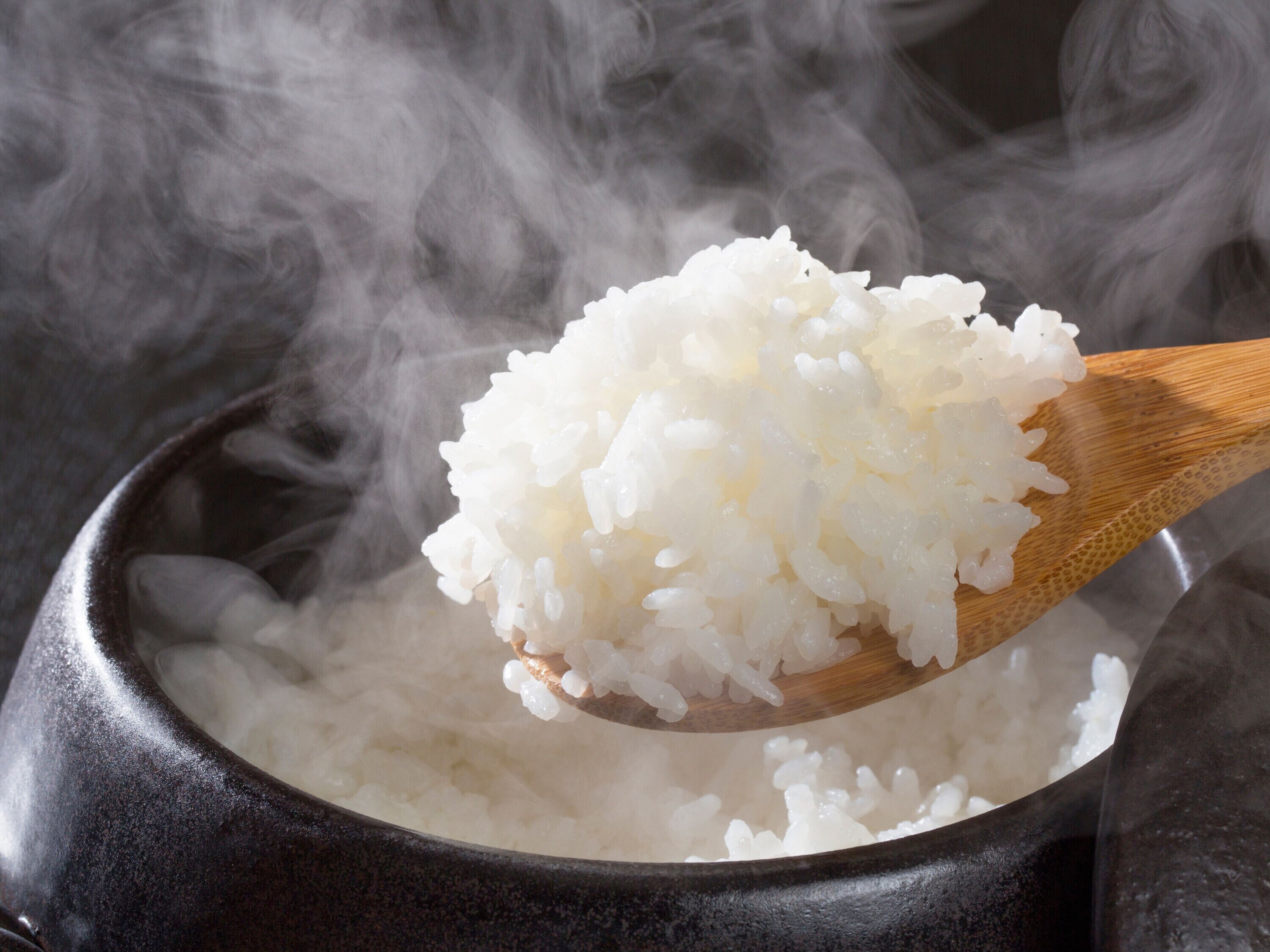 A wooden spoon filled with white rice being lifted from a pot, surrounded by steam.