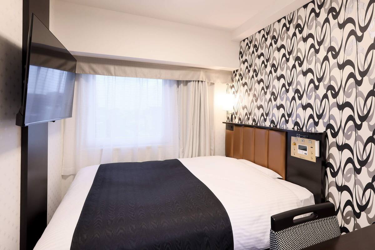 Une chambre d’hôtel comprenant un lit, une télévision, une tête de lit avec des commandes, une fenêtre avec des rideaux et un mur décoré d’un motif.