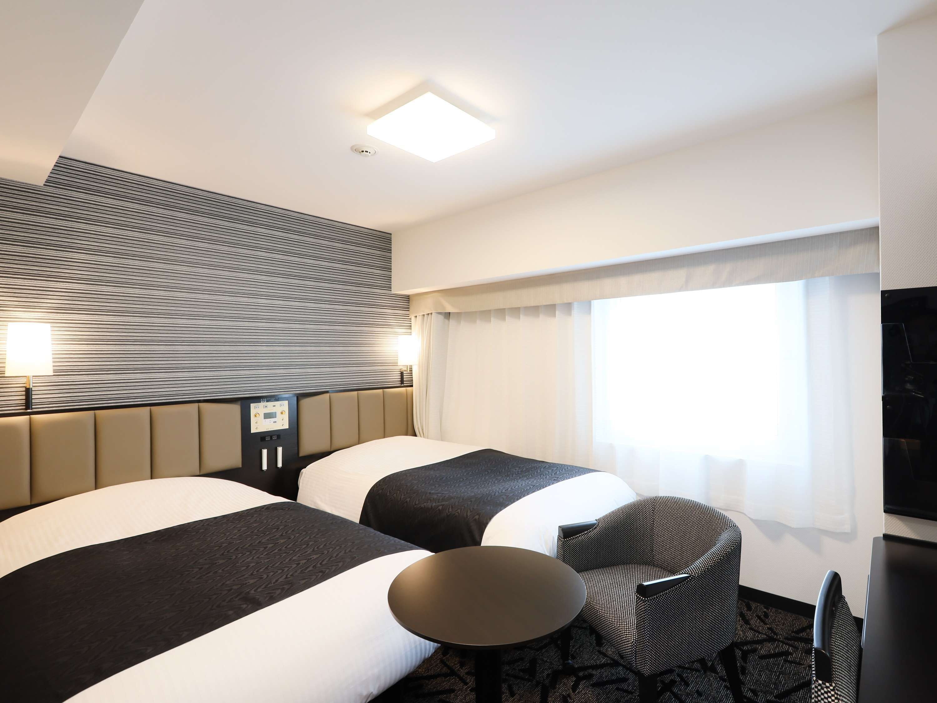Habitación de hotel con dos camas, una mesita, una silla, televisor y ventana con cortinas.