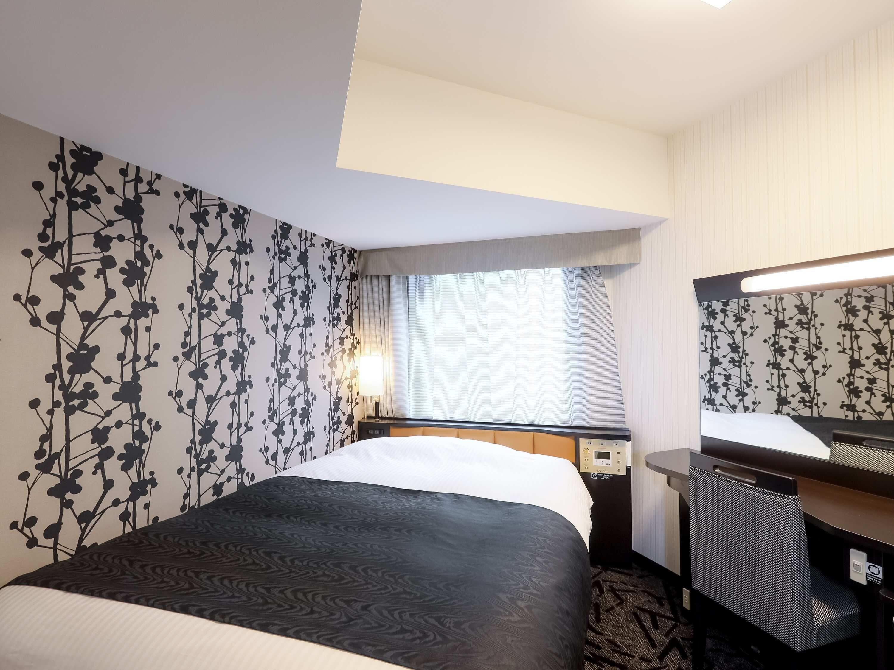 A hotel room with a bed, a desk, a chair, a window with curtains, and a wall with a floral pattern.