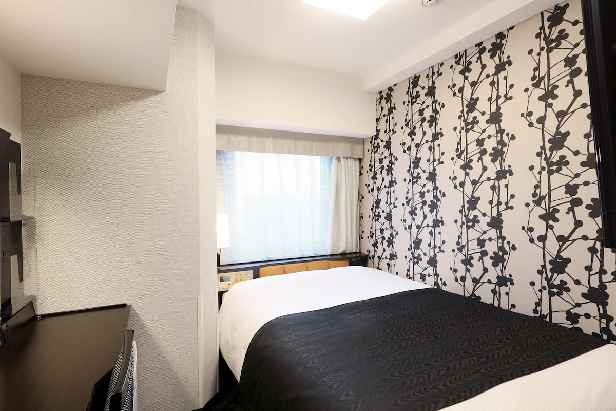 Une chambre d’hôtel avec un lit, un bureau, une fenêtre avec des rideaux et un mur orné d’un motif floral.