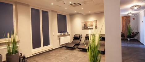 A modern spa room with a massage table, two large potted plants, a radiator, and a wall-mounted artwork.