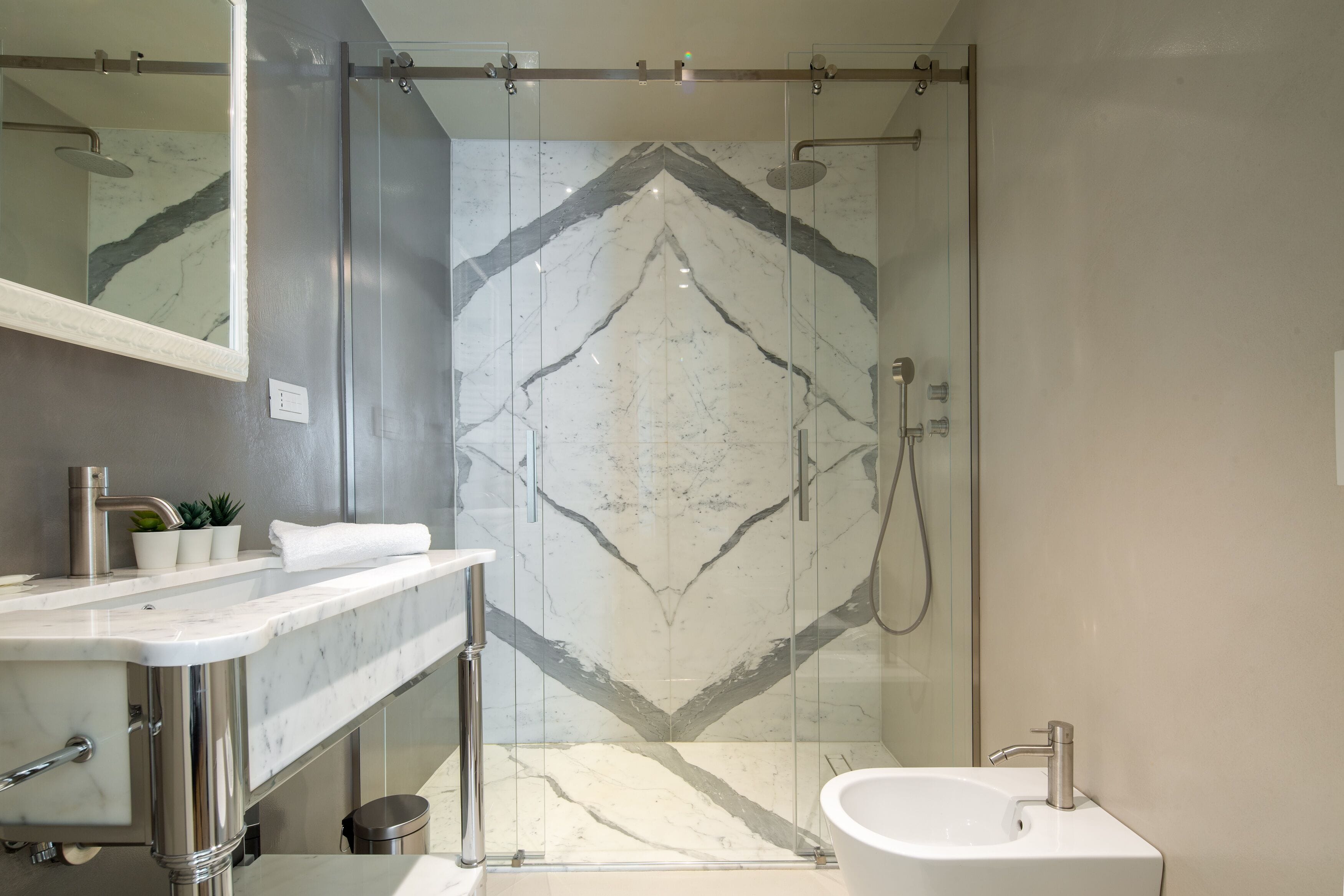 Ein modernes Badezimmer mit Duschkabine aus Glas, einer an der Wand gefliesten Duschkabine mit Marmor und einem weißen Waschbecken mit Marmorplatte.