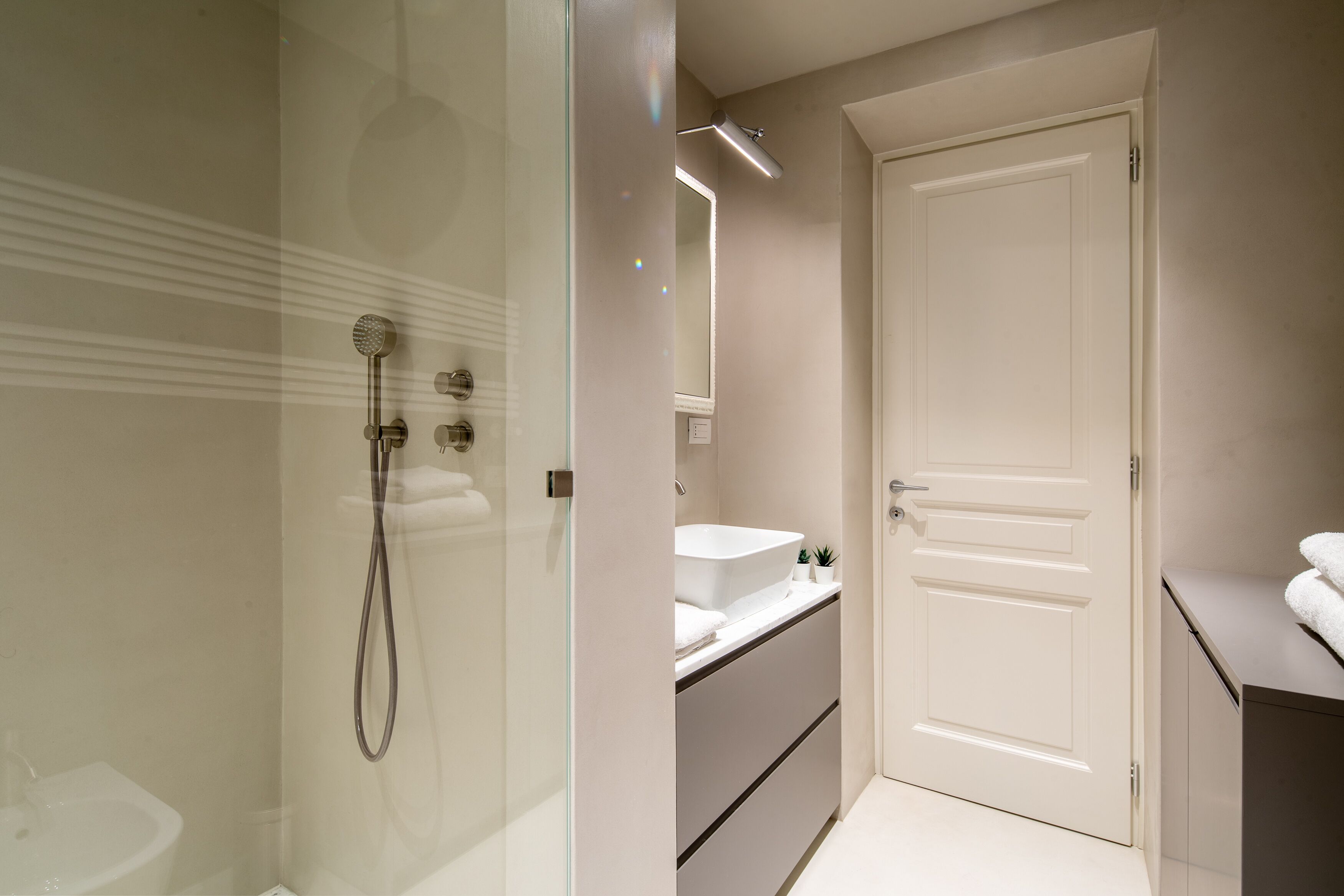 A modern bathroom with a glass-enclosed shower, a white sink, and a door with a mirror above it.