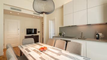 A modern kitchen with a dining area, featuring a marble table, grey upholstered chairs, a black pendant light, and a built-in TV.
