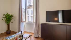 A modern living room with a large window, a potted plant, a glass coffee table with books and a bottle, and a flat-screen TV on a dark cabinet. - Florence Luxury Apartment al Duomo by Mmega (Florence)