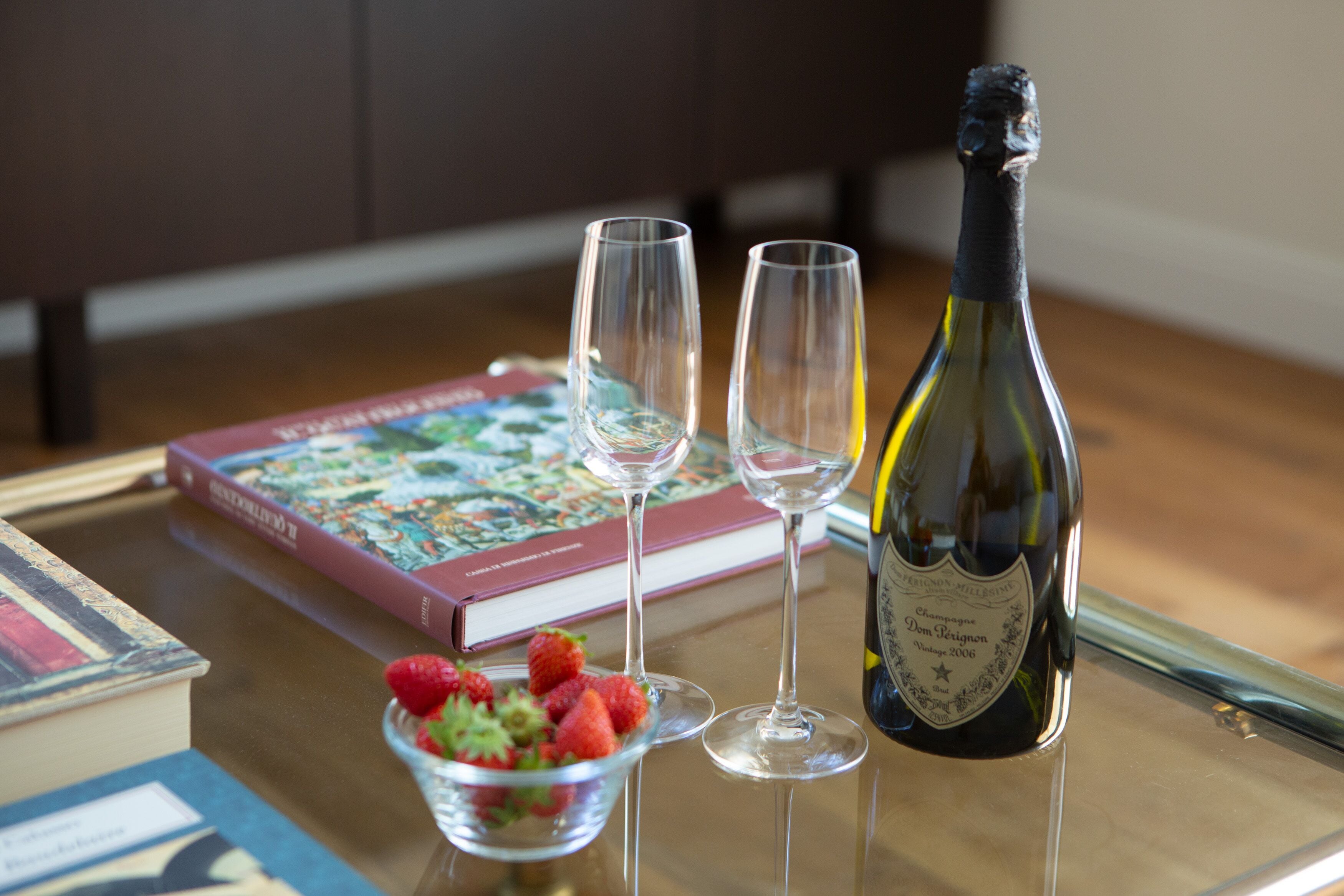 A table with a bottle of champagne, two empty wine glasses, a bowl of strawberries, and a book.