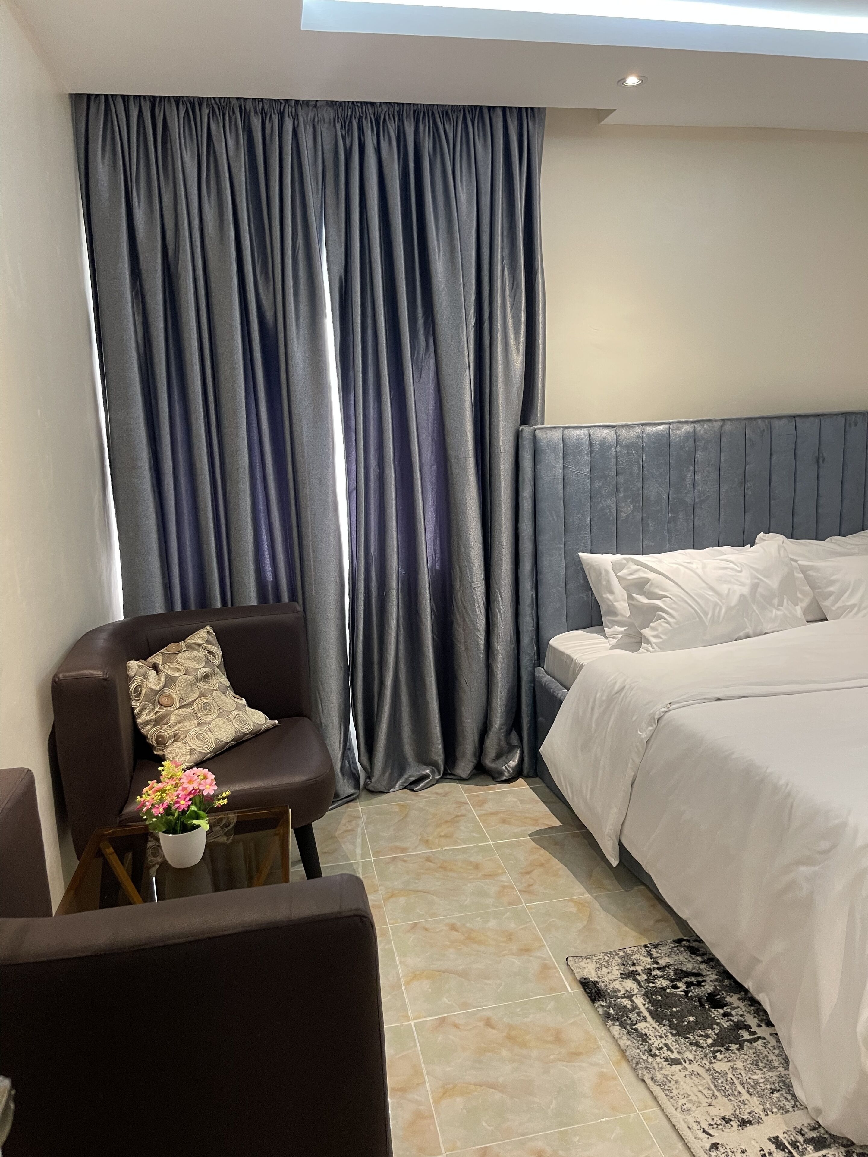 Une chambre d’hôtel moderne, équipée d’un lit, d’une chaise, d’une petite table ornée d’un vase de fleurs et d’une grande fenêtre avec des rideaux.