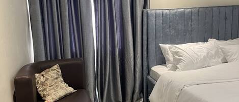 Une chambre d’hôtel moderne, équipée d’un lit, d’une chaise, d’une petite table ornée d’un vase de fleurs et d’une grande fenêtre avec des rideaux.