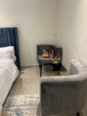 A bedroom with a bed, a grey armchair, a small table with a vase of flowers, and a dark blue headboard.