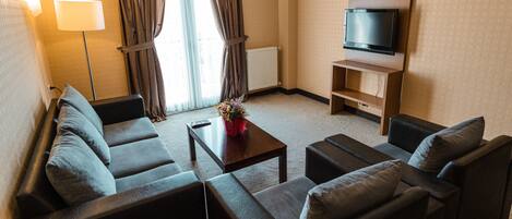 A hotel room with a sofa set, a coffee table, a TV on a stand, and a window with curtains.