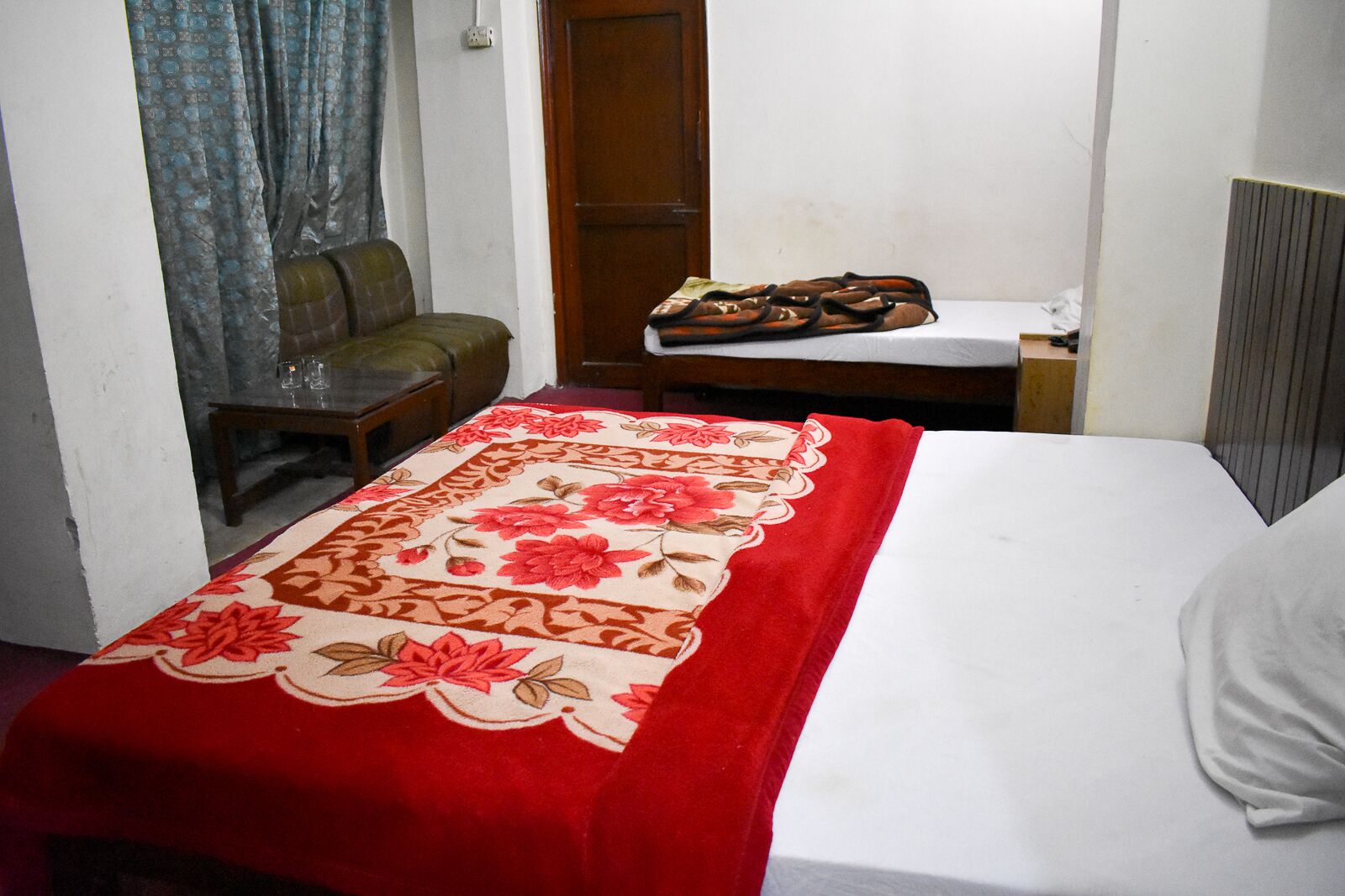 A small, simple room with a bed, a wooden chair, a table with shoes, and a curtain.