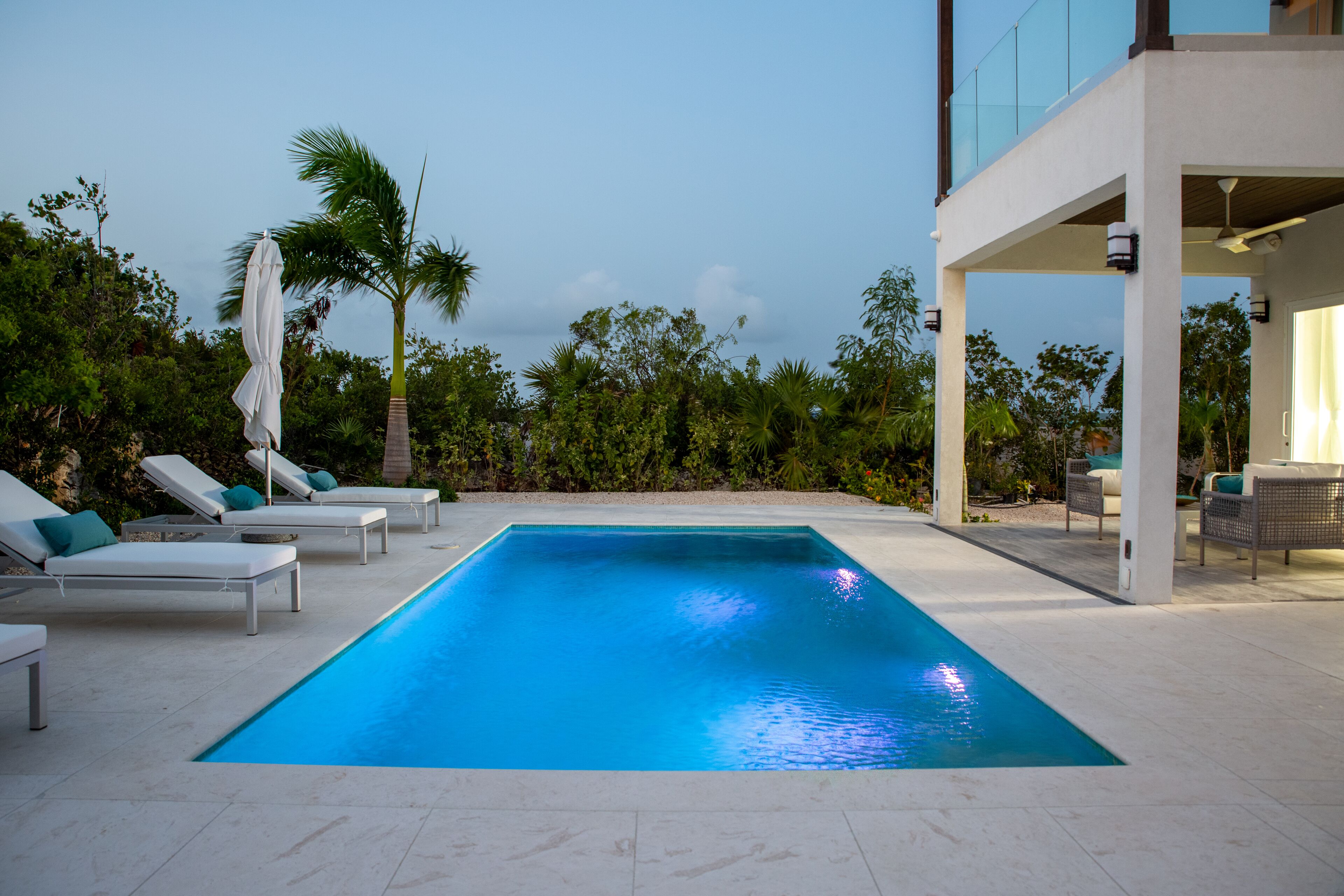 Una piscina rectangular con sillas de descanso y sombrillas, rodeada de vegetación y arquitectura moderna.