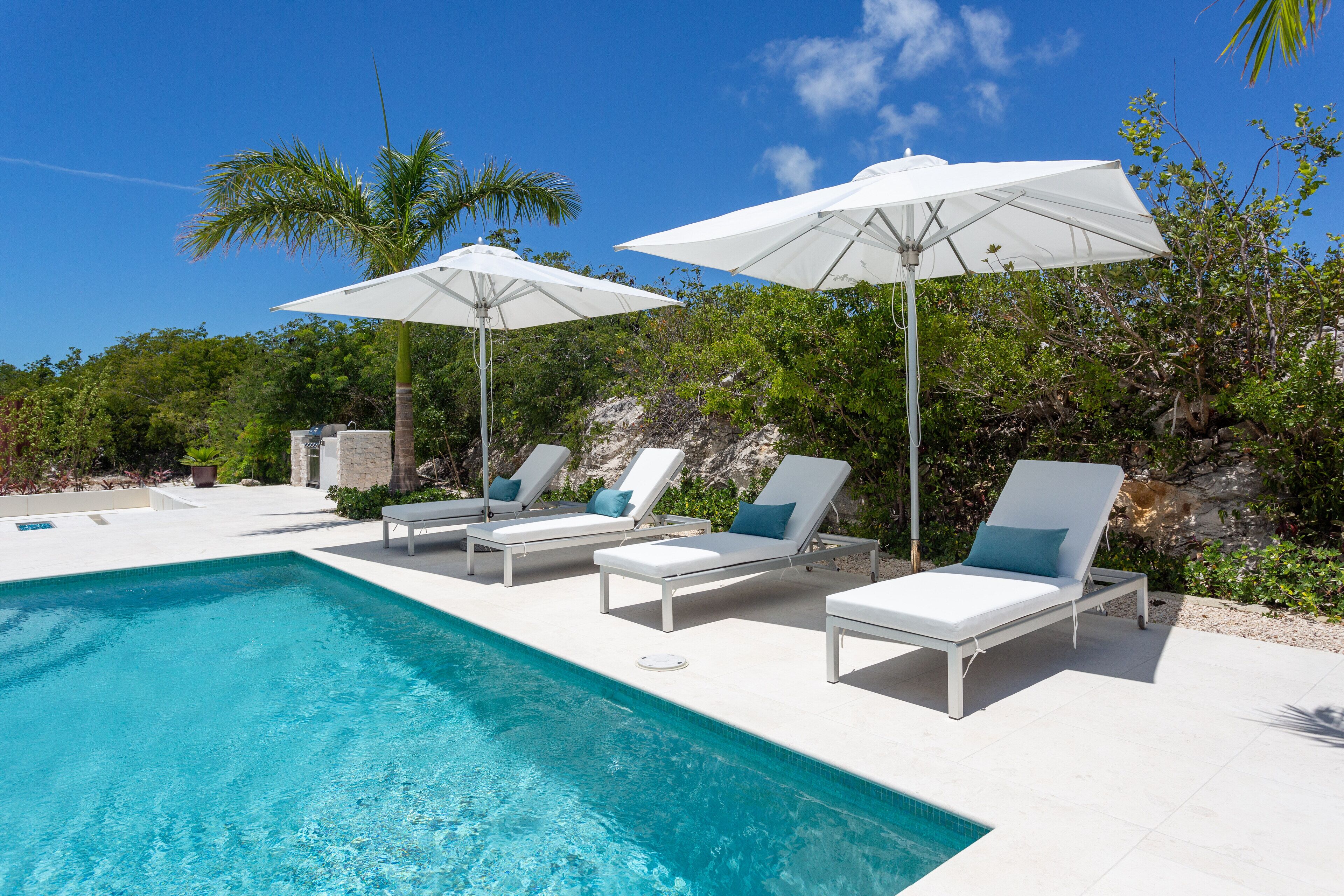 A swimming pool with white lounge chairs and umbrellas, surrounded by greenery and a clear sky.