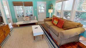 A cozy living room with a sofa, a bench, and a coffee table. There is a door with a window, a lamp, and a view of the outdoors. - Classic Beach Cottage~Surf Shack (Folly Beach)