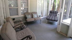 A covered porch with beige cushioned seating, a metal chair, and a glass door leading outside. - Classic Beach Cottage~Surf Shack (Folly Beach)