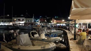 A marina at night with boats docked, a café with outdoor seating, and buildings in the background. - Bluetel Lixouri, Greece (Kefalonia)