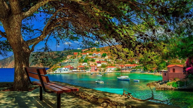 A bench by a clear blue sea with a tree overhead, overlooking a coastal village with colorful buildings. - Bluetel Lixouri, Greece (Kefalonia)