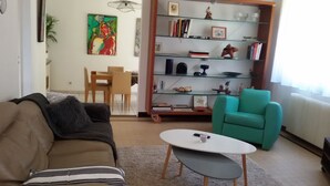 A living room with a brown sofa, a teal armchair, a small white table, and a bookshelf filled with books and decorative items.