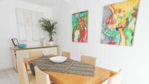 A dining area with a wooden table, beige chairs, a patterned table runner, and two colorful paintings on the wall.