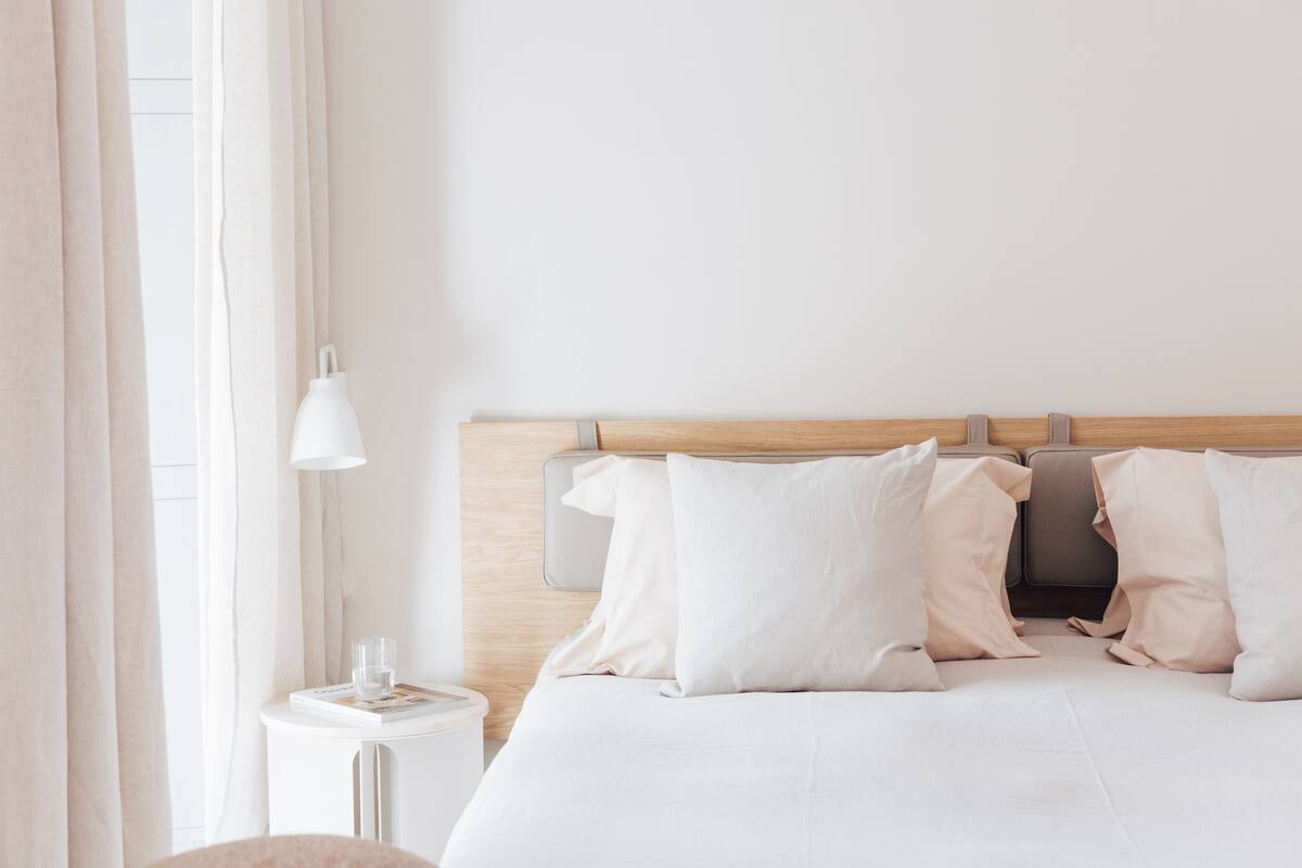 A neatly made bed with white linens and pillows, a bedside table with a lamp, and a window with curtains.