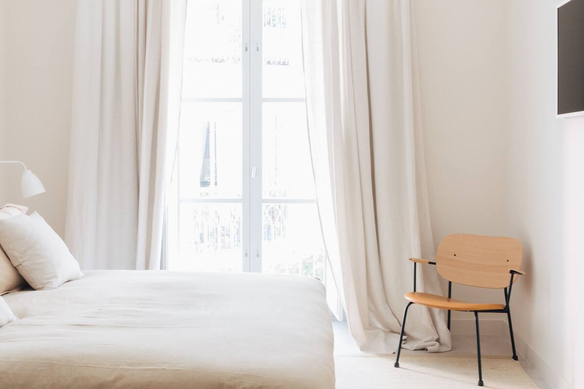 A bedroom with a bed, a chair, a window with curtains, and a wall-mounted air conditioning unit.