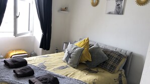 A bedroom with a bed, pillows, a blanket, a wall-mounted shelf with a figurine, and a window with curtains. - Captivating 2-bed Apartment in Isle of Bute (Rothesay)