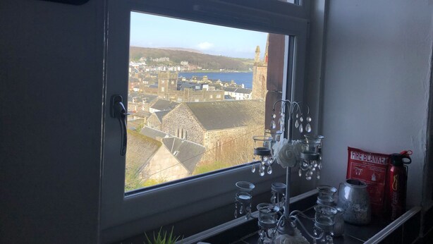 A window with a view of a town, a shelf with glassware, and a fire blanket. - Captivating 2-bed Apartment in Isle of Bute (Rothesay)