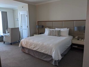 Ein Hotelzimmer mit einem großen Bett, zwei Nachttischen mit Lampen, einem Sessel und einer Tür zu einem weiteren Raum.