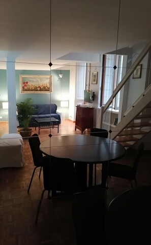 Uma sala de jantar com uma mesa redonda e cadeiras, um sofá, uma planta em vaso e uma escada que sobe.