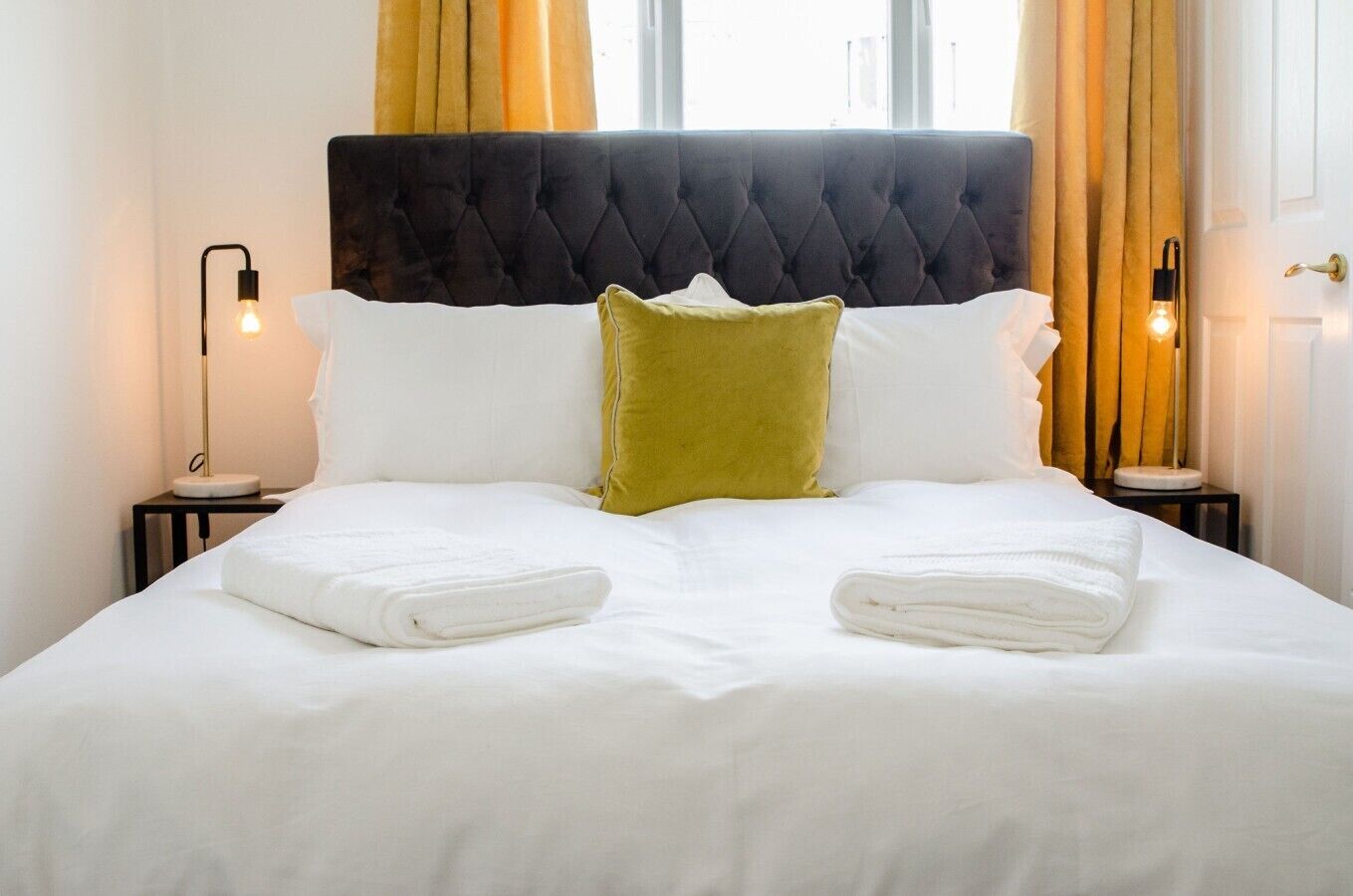 Un lit bien fait, avec du linge de lit blanc et deux oreillers, un vert et un blanc, dans une chambre dotée d’une tête de lit capitonnée et de deux lampes de chevet.