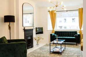 A living room with a green sofa, a fireplace, a flat-screen TV, and a modern chandelier.