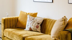 En stue med en sennepsgul sofa, en vægmonteret lampe og to indrammede kunstværker.