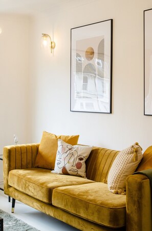 A living room with a mustard-colored sofa, a wall-mounted light fixture, and two framed artworks.