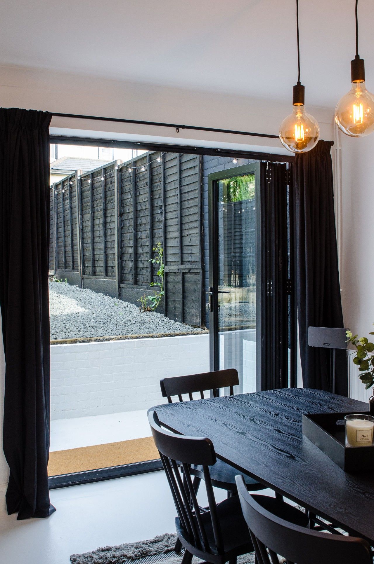 A modern dining area with a dark wooden table and chairs, a candle on the table, and a view through a glass door to an outdoor area with a gravel path and greenery.